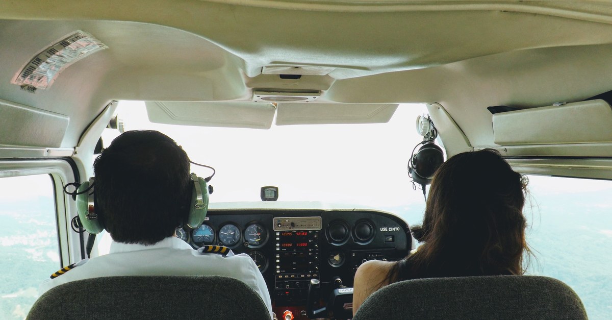 View from behind in a small-plane cockpit: an experienced pilot and a newcomer sit side by side at the controls, flying forward over open country.