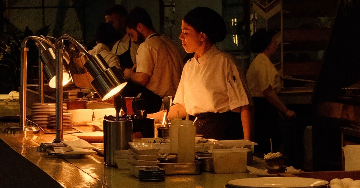 A chef at the pass under warm kitchen lamps at night, line cooks working behind and plates stacked for service.