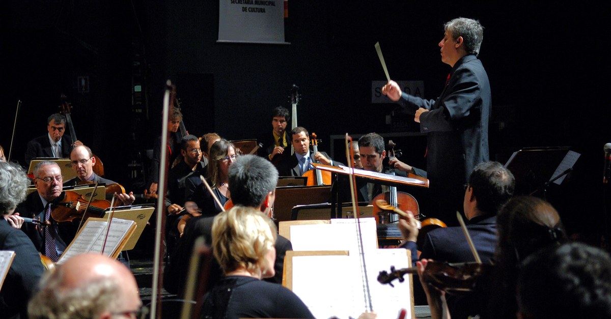 A conductor with baton raised leads a full orchestra mid-performance, the players holding to his direction.