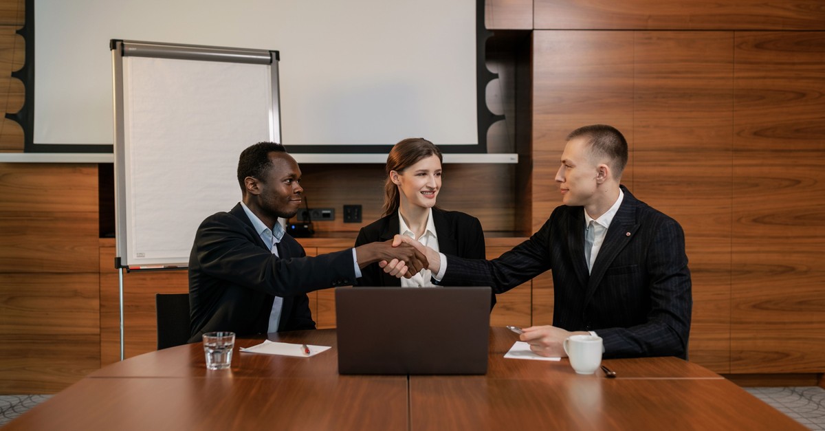 A woman seated between two businessmen at a conference table as they shake hands over an open laptop, the negotiator in the middle who got both sides to yes.