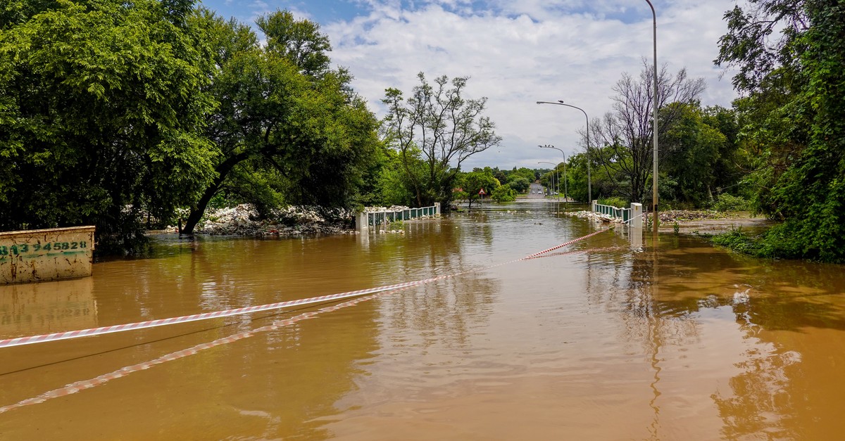 A residential street completely submerged by muddy brown floodwater, with a thin red-and-white barrier tape strung across it as the only marker: a sign standing in for the fence that was needed.