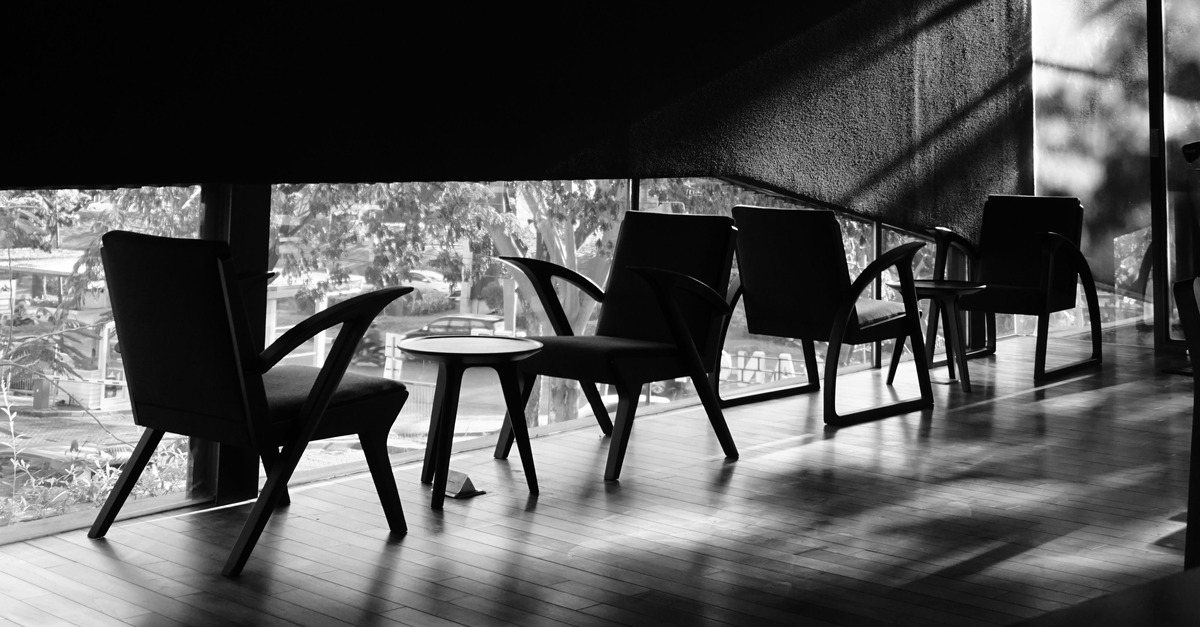 Empty chairs and a low table silhouetted in a dark room, with daylight coming through the window behind them.