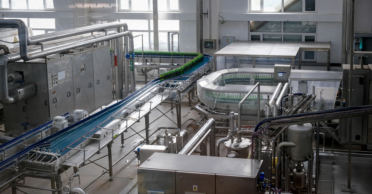 A long, empty conveyor belt curving through a bottling plant, with bottles bunched tightly together on the line at the far end.